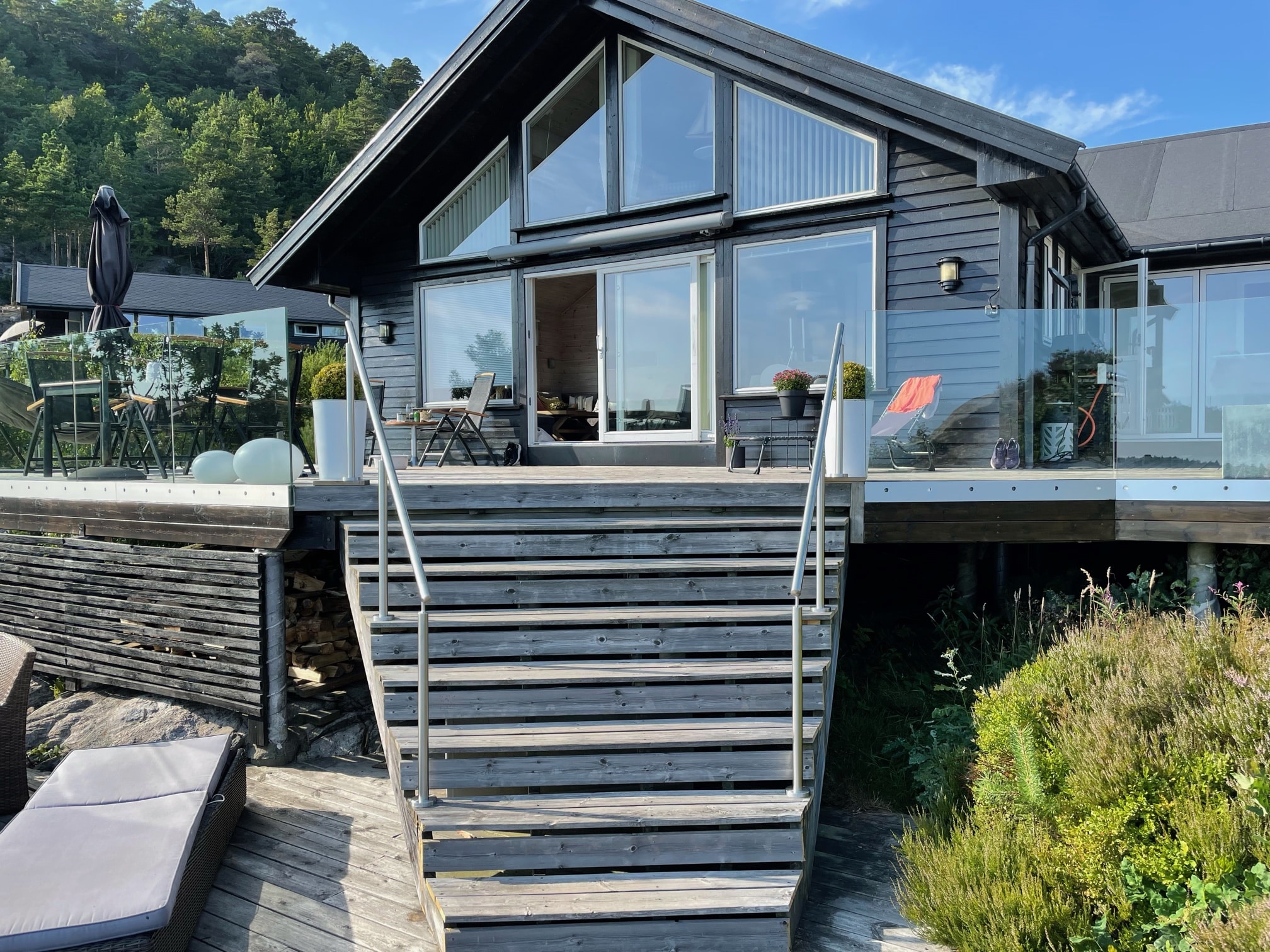 Stainless steel wire handrails installed on wide wooden stairs leading to a black cabin-style house with glass balustrades and hillside views.